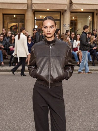 Woman in a brown leather bomber jacket and black trousers standing in a busy street style crowd