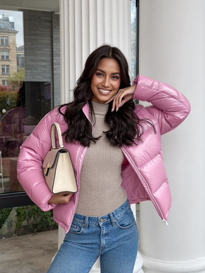 Woman smiling in a glossy pink puffer jacket layered over a taupe knit top and blue jeans