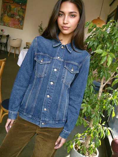 Woman in a blue denim jacket and brown corduroy trousers beside a potted plant